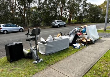 A large pile of rubbish set out on the curb for council collection in a suburban neighbourhood. The heap includes oversized items like flattened cardboard boxes, a worn couch with pillows, several mattresses stacked atop one another